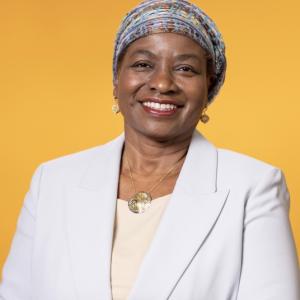 portrait of smiling woman in business wear, and a headwrap against a deep yellow background 