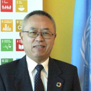 portrait of man in suit, tie, and glasses with an SDG banner and UN flag in background