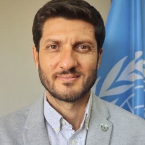 portrait of man with brown hair and a beard, with a slight smile in shirt and blazer with a UN pin and UN flag inthe background