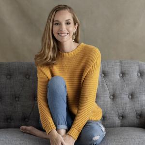 woman with blonde hair in sweater and jeans on a nice couch smiling for a photo